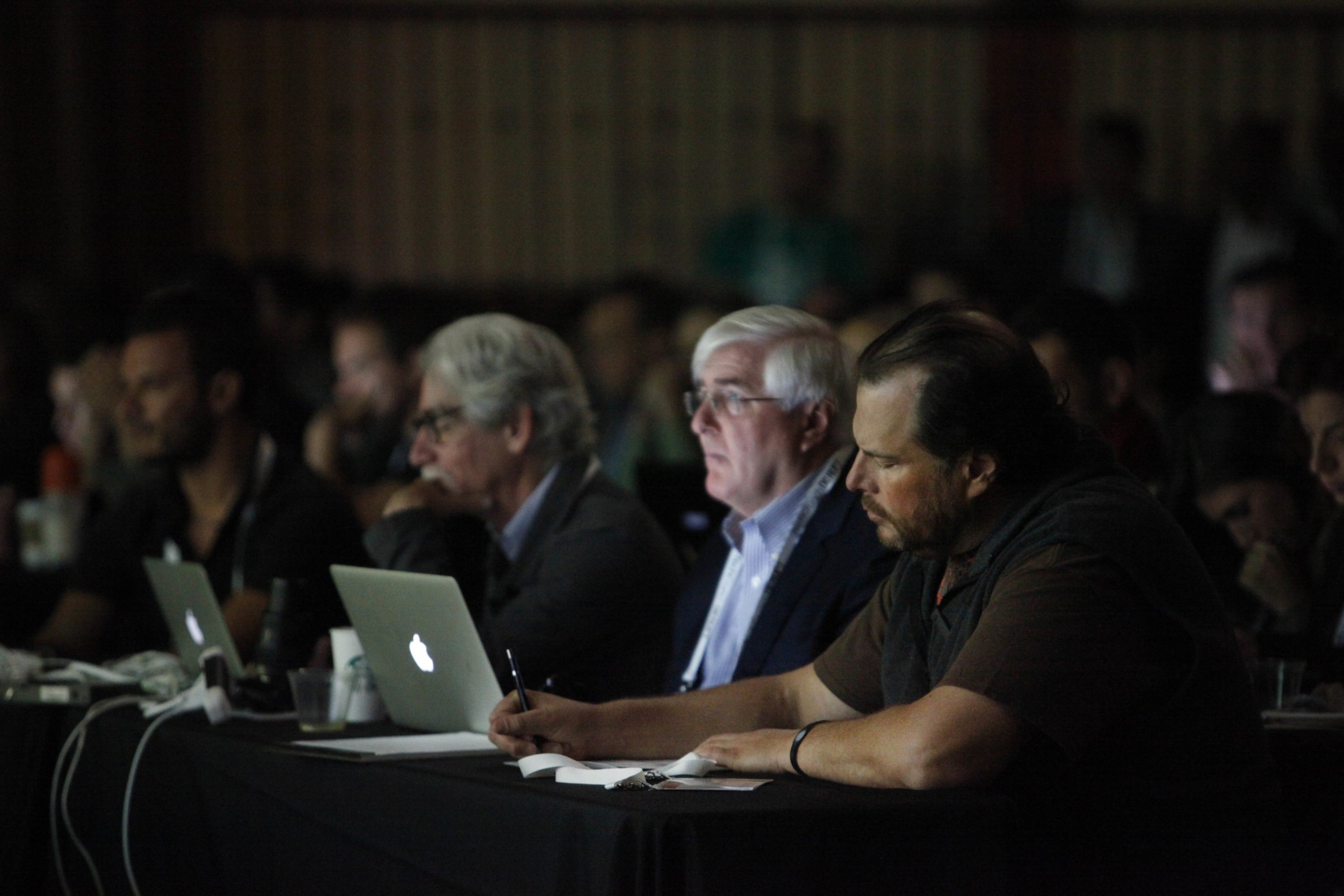 Several men sit at a long table with laptops, writing notes and focused on a presentation in a dimly lit room.