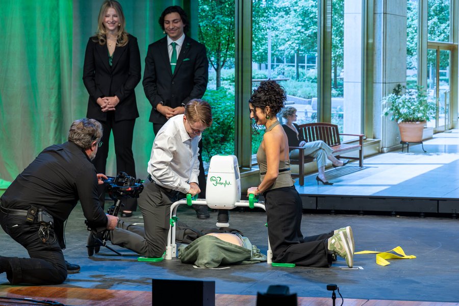 Two kneeling students demonstrate an automated CPR device. Two standing students watch, and a photographer kneels for a close-up shot.