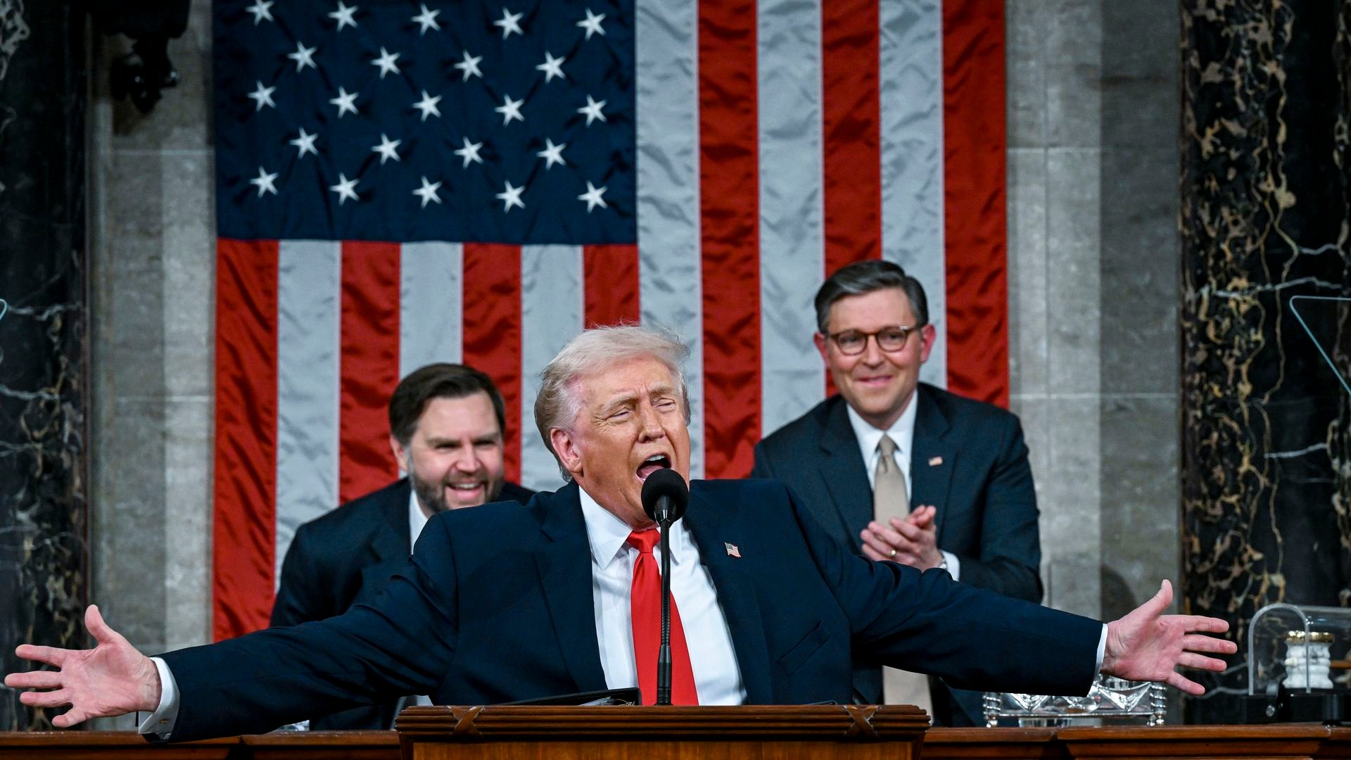 Trump speaking into a microphone with his arms spread wide wearing a navy suit jacket with an American flag pin, white collared shirt and red tie while Speaker Mike Johnson and Vice President JD Vance, standing behind the president, clap and smile