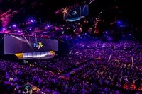 Wide view of a large audience in a purple-lit arena looking at a main stage screen displaying 