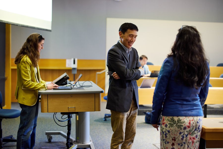Three people standing in a lecture hall. A woman at a lectern works on her laptop while two others converse in the foreground.