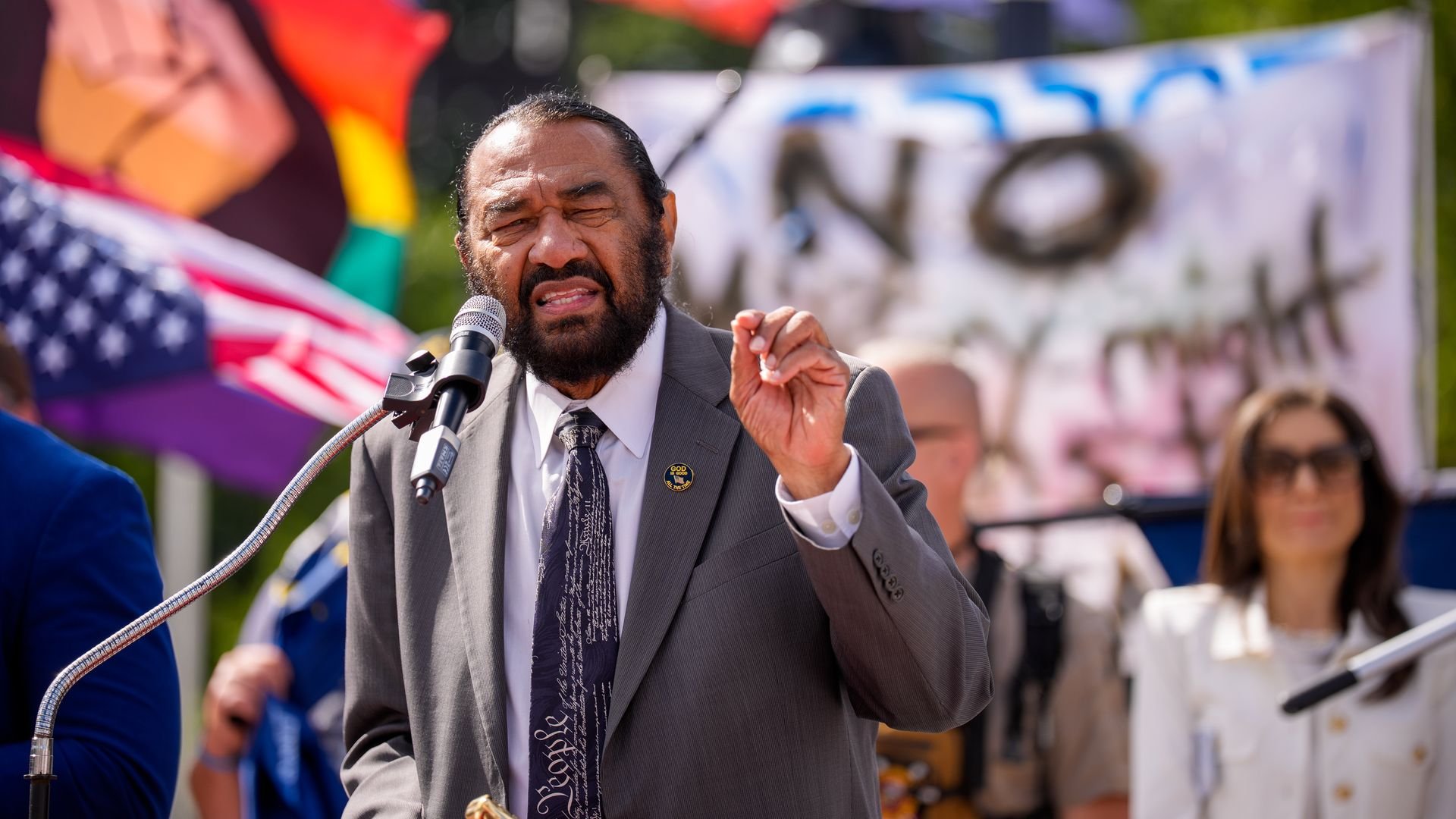A man in a gray suit and a Constitution-themed tie speaks passionately at a microphone outdoors, with colorful flags and a blurred protest sign in the background.