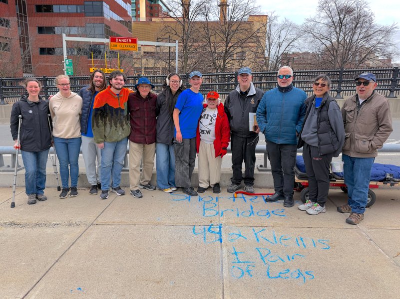 12 people stand together at the end of a bridge with the writing, “Shortfellow Bridge - 442 Kleins + pair of legs” in chalk on the sidewalk.