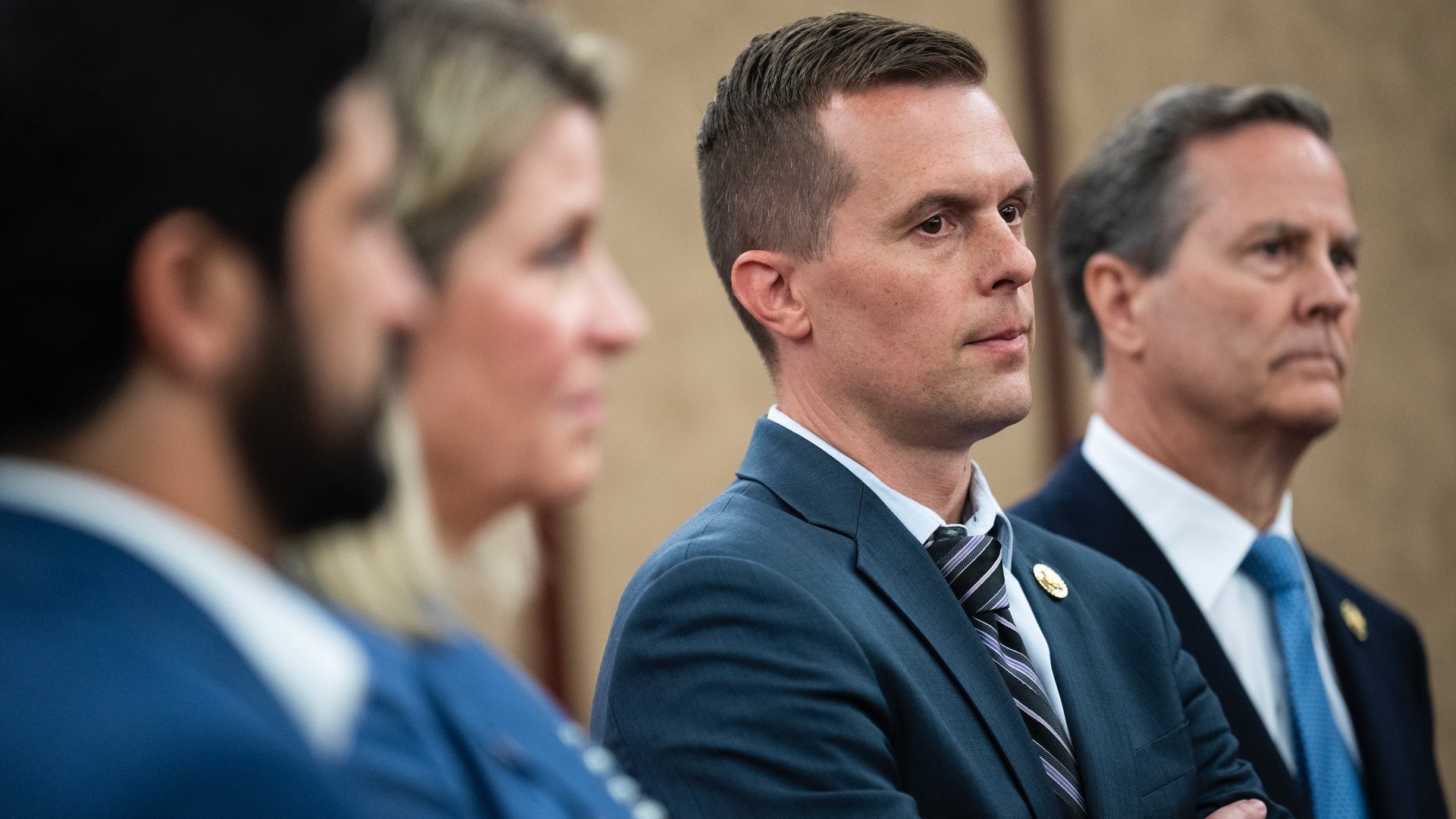 Four people in business attire standing in a line, two men in focus wearing suits and ties, one with arms crossed, neutral background.