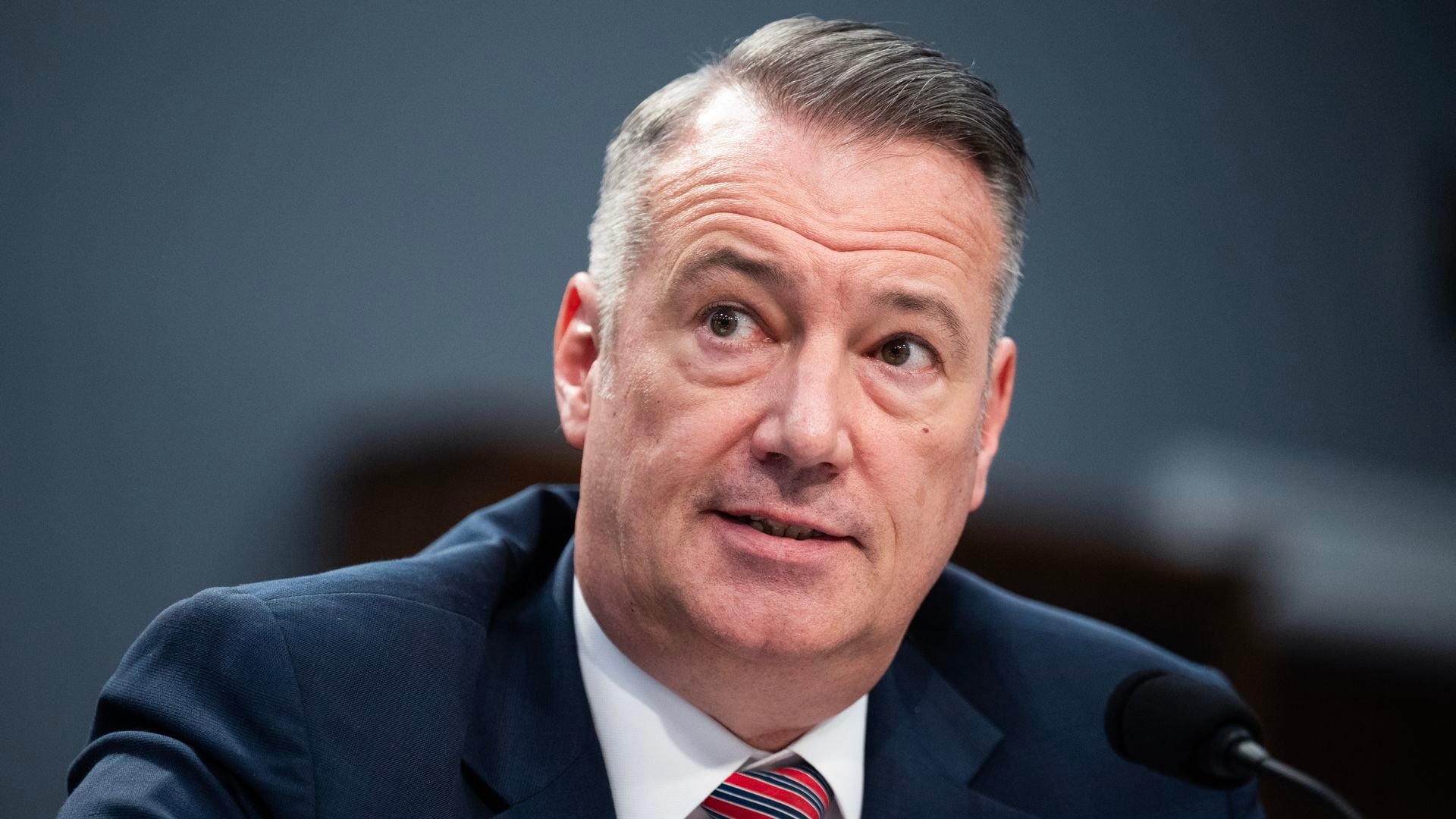A close-up of a middle-aged man in a navy suit and striped red-blue tie, with a small American flag pin, speaking into a microphone; gray hair, looking to the side, blue-gray background.