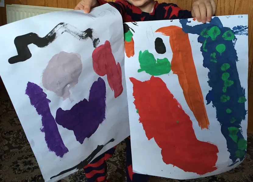 A child holding up two large abstract paintings in bold poster-paint colours