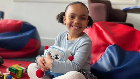 BBC Picture of a young girl hugging Gabbo an AI-powered toy. She is sat, smiling among soft seating and other toys.