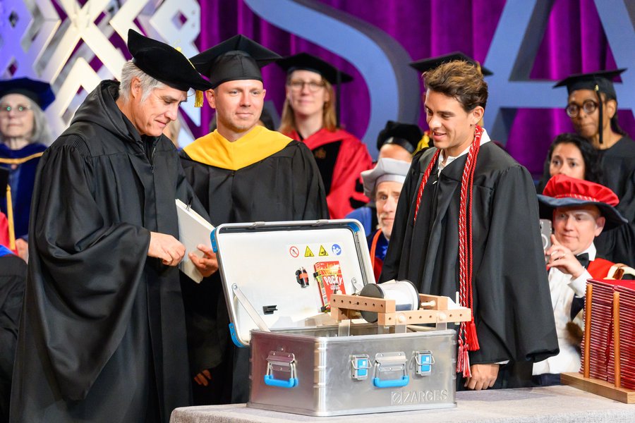 Alejandro Aravena opens a large silver toolbox filled with handmade objects while Oliver Moldow and Skylar Tibbits stand beside him on stage in academic regalia. Faculty members are seated behind them.