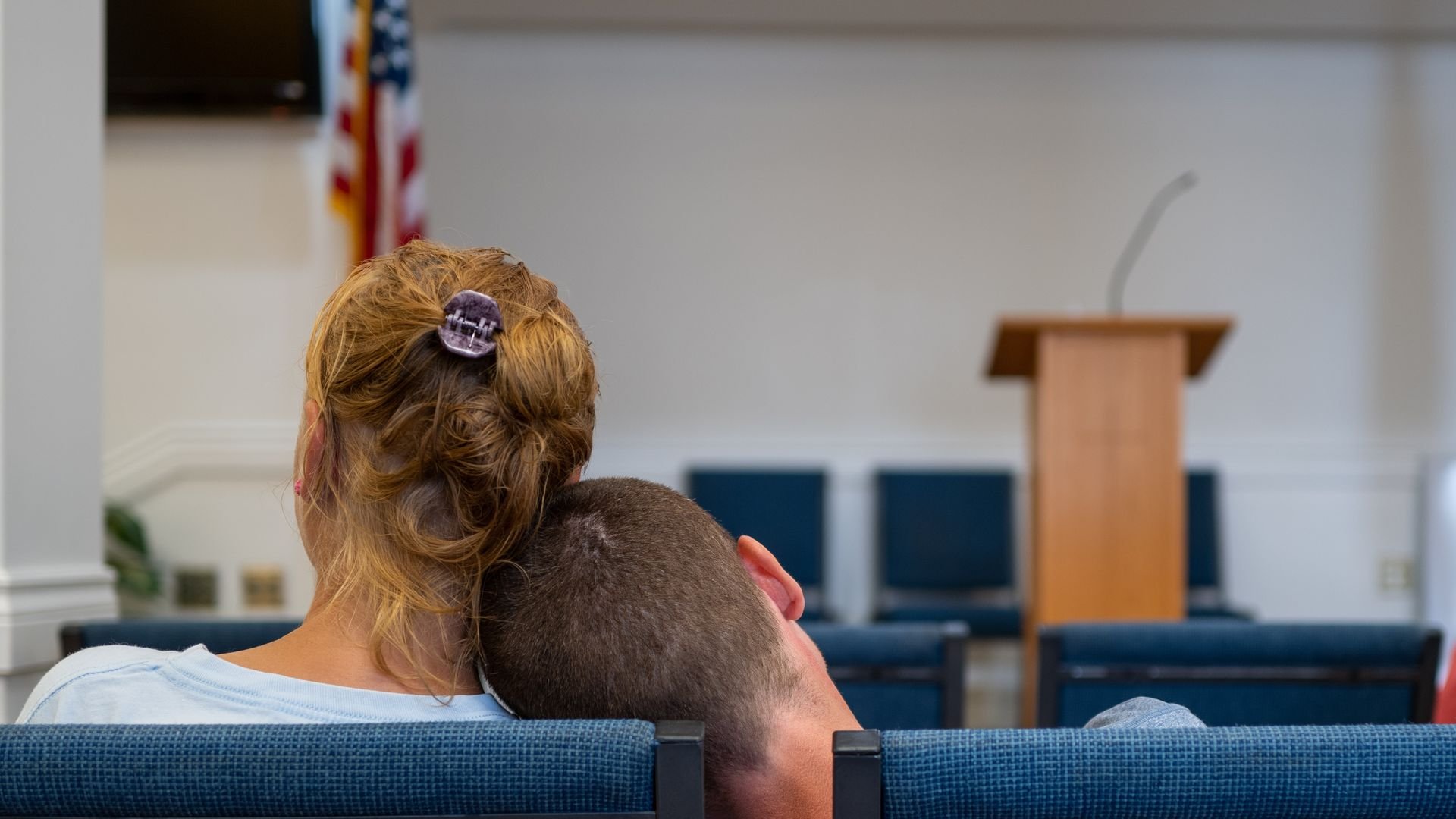 Two people sitting on blue chairs in a room with an American flag, a podium with a microphone, and empty chairs in the background. One person's head rests on the other's shoulder.