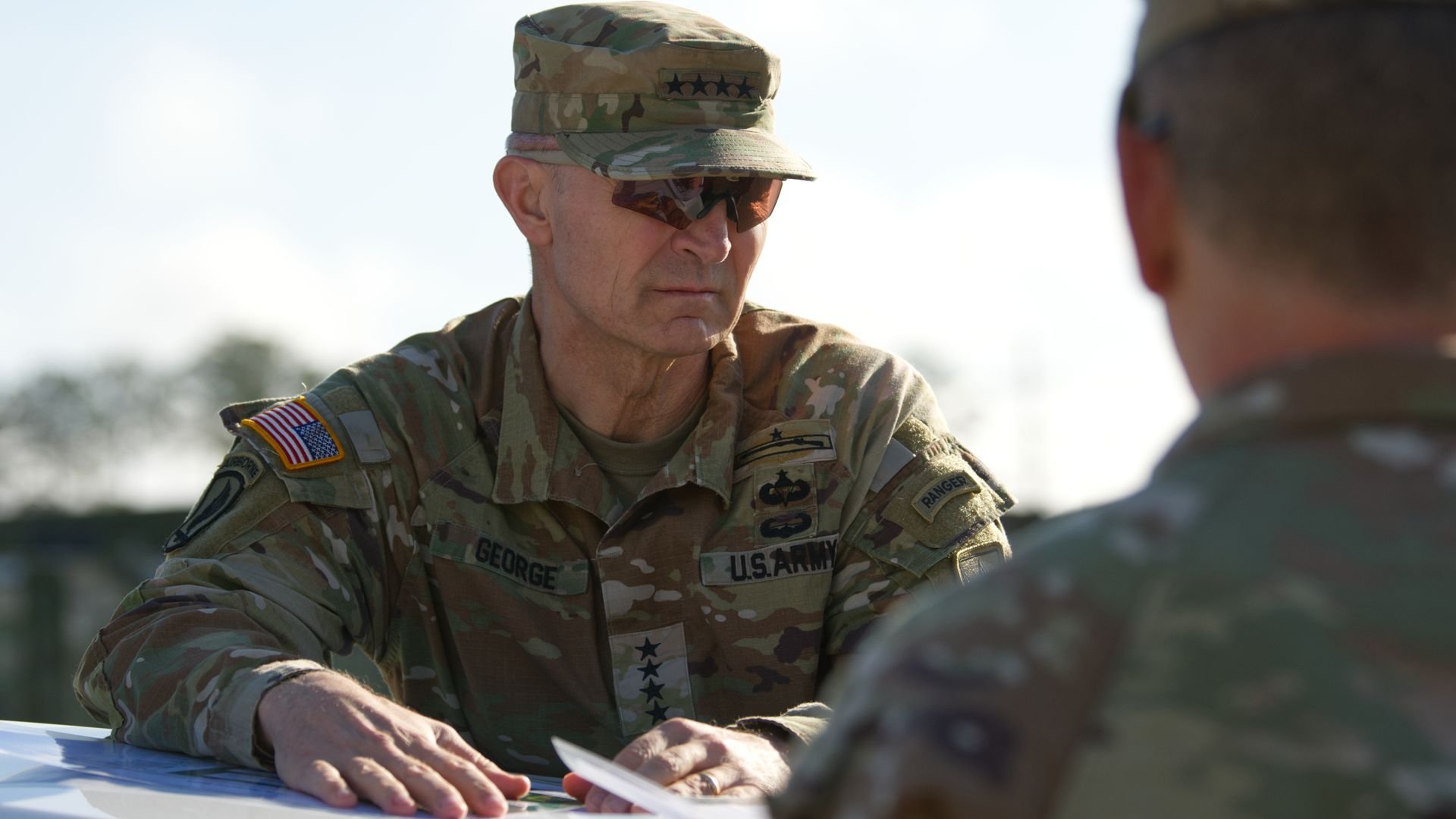 Two U.S. Army officials in camouflage talk outdoors. The man facing the camera wears a camo cap and sunglasses. Another man, in the foreground, has his back to the camera.