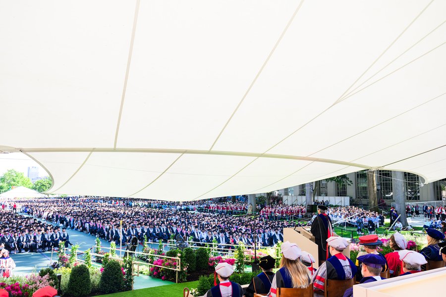 Rows of graduates sit on Killian Court; in the foreground a speaker stands at the podium under a large canopy.