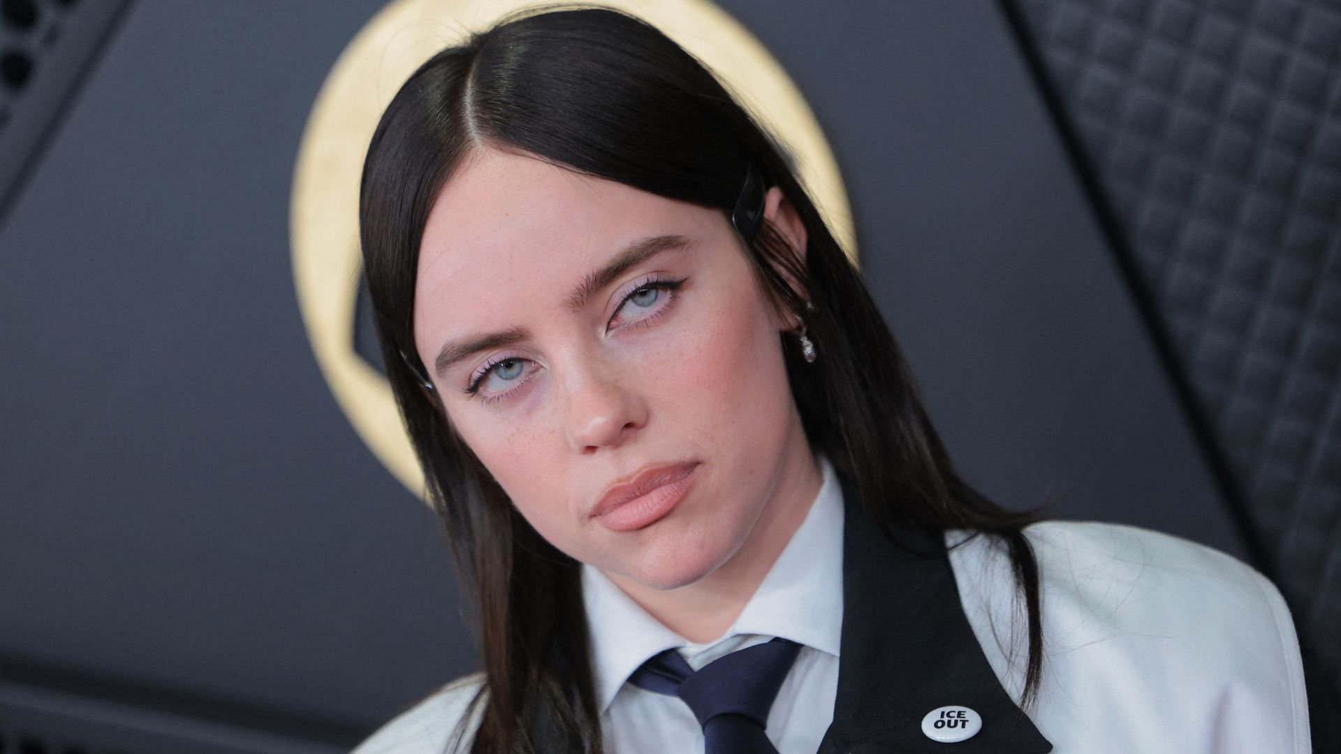 Close-up of a woman, singer songwriter Billie Eilish, with blue eyes, brown hair, wearing a white shirt, dark tie, and jacket with an 