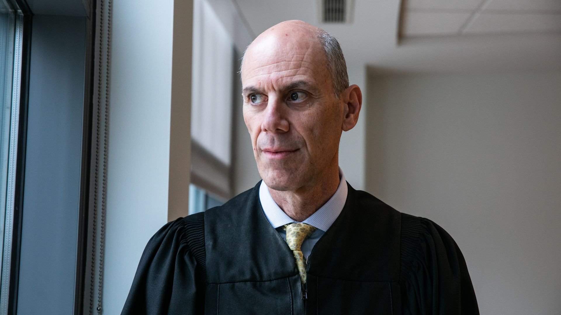 Judge Boasberg — wearing his black robe, a light-colored collared shirt and a yellow tie — looks out a window.
