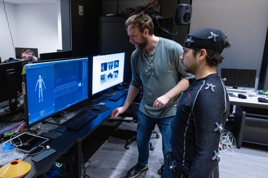 Two people standing at a desk work to pair a motion capture suit with the avatar on screen.
