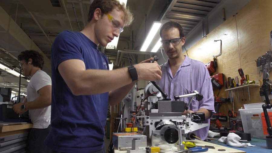 Two students wearing safety glasses in a lab examine a lathe, while a third works on something else in the background
