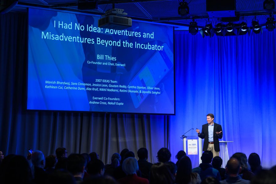 Bill Thies speaks on an indoor stage under a screen showing his keynote title: 
