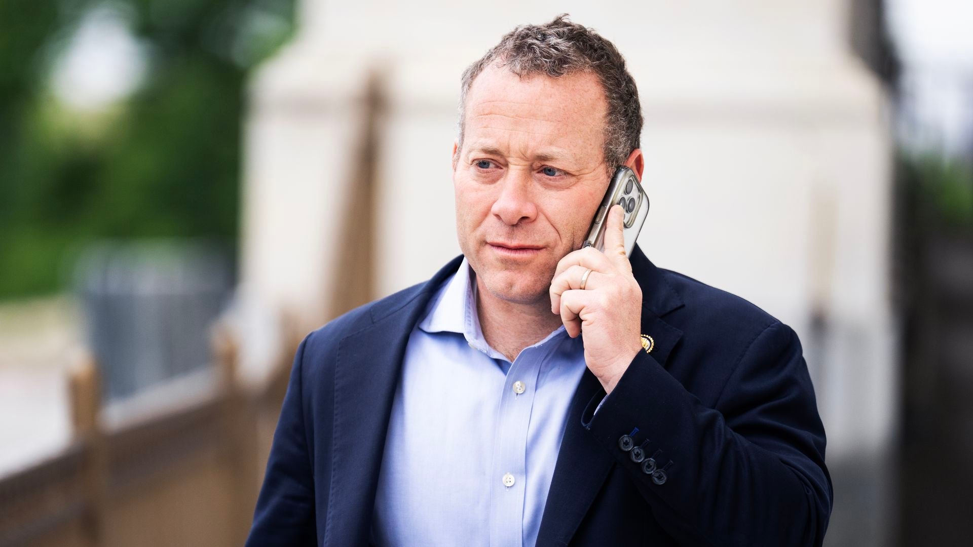 Man with curly hair, wearing a navy blazer and light blue shirt, holding a phone to his ear, standing outdoors near a railing and blurred background.