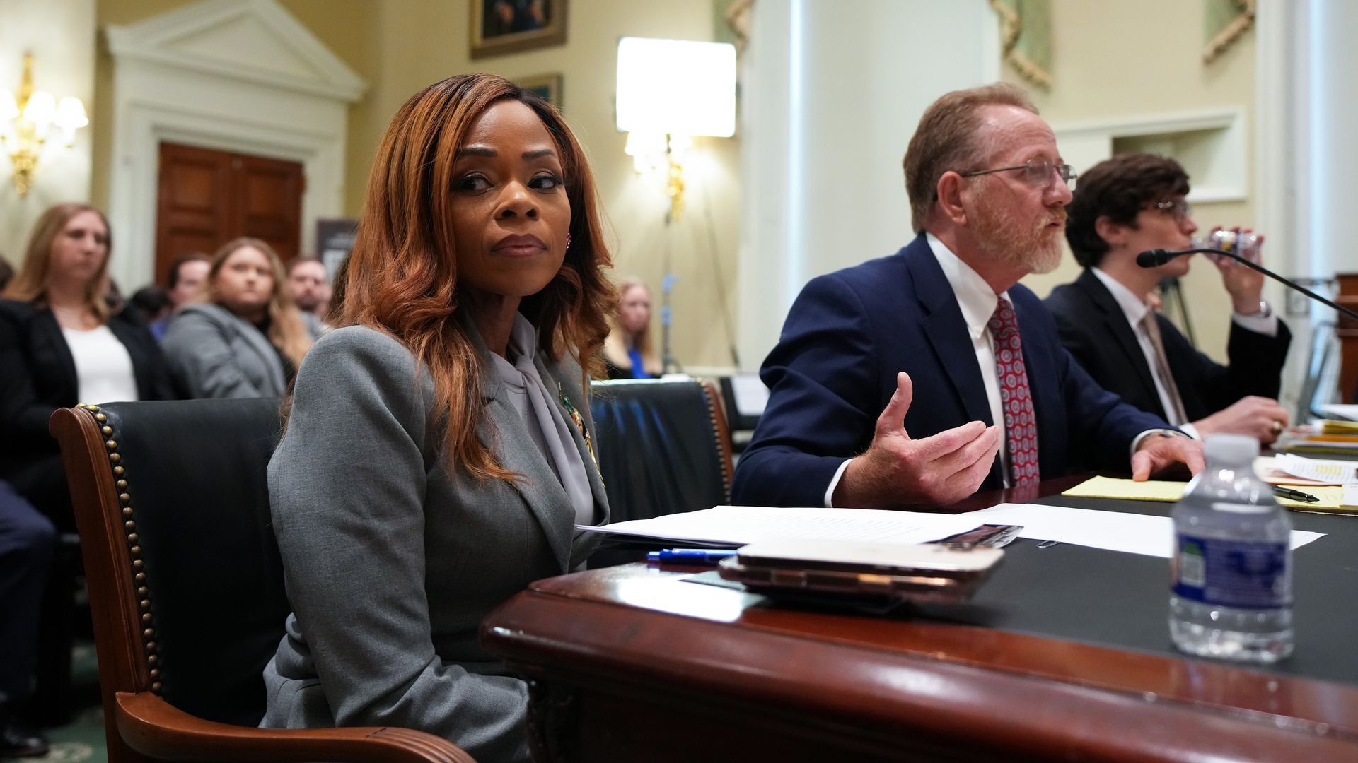 Woman with brown hair in a gray blazer sits at a dark wood desk in a formal meeting room; two suited men speak at a panel in the background, with papers and a water bottle on the desk.