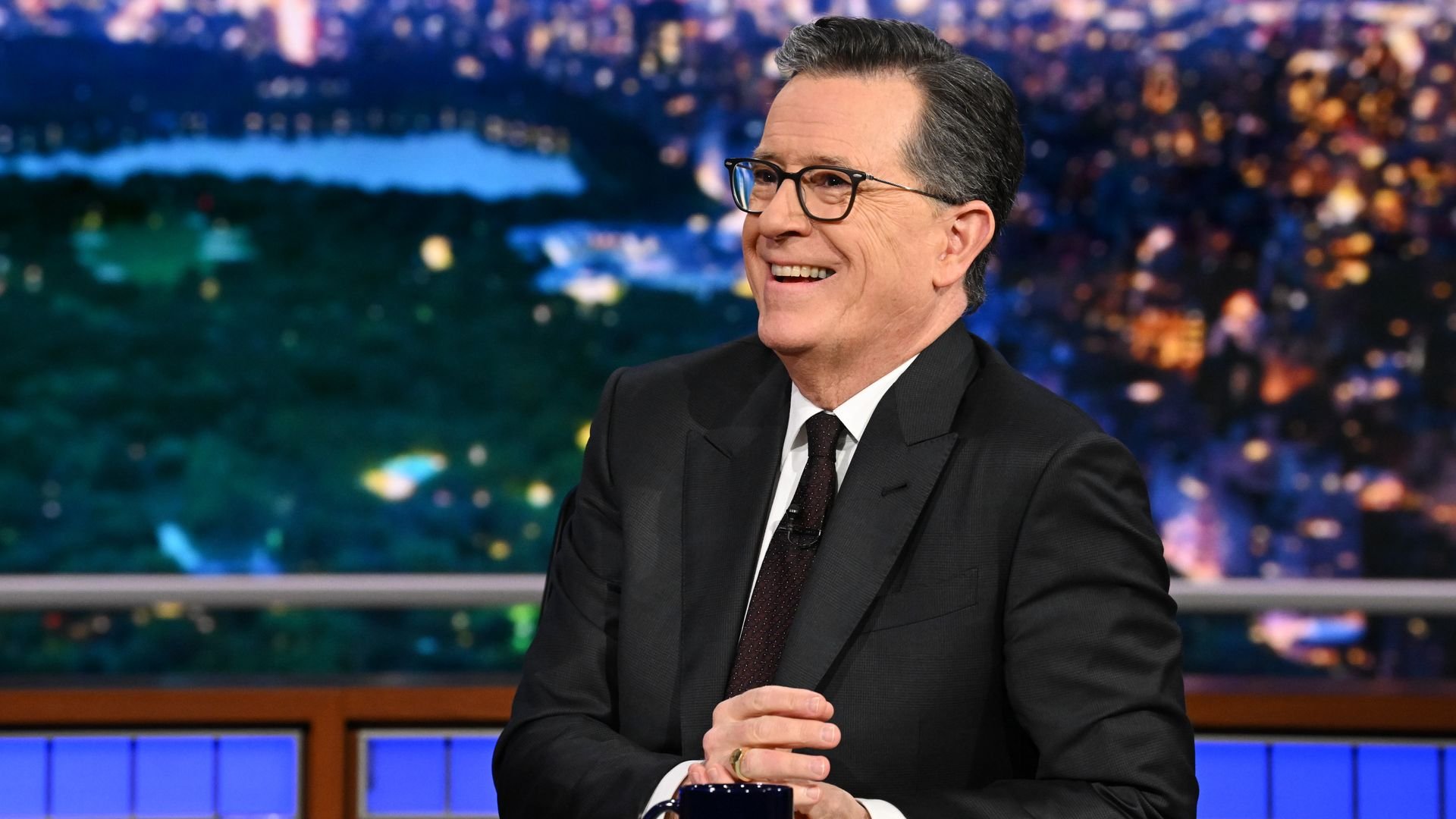 Stephen Colbert — wearing a dark suit and tie with a white collared shirt and glasses — smiles from behind his desk on stage. A black mug with red lettering sits on his desk.