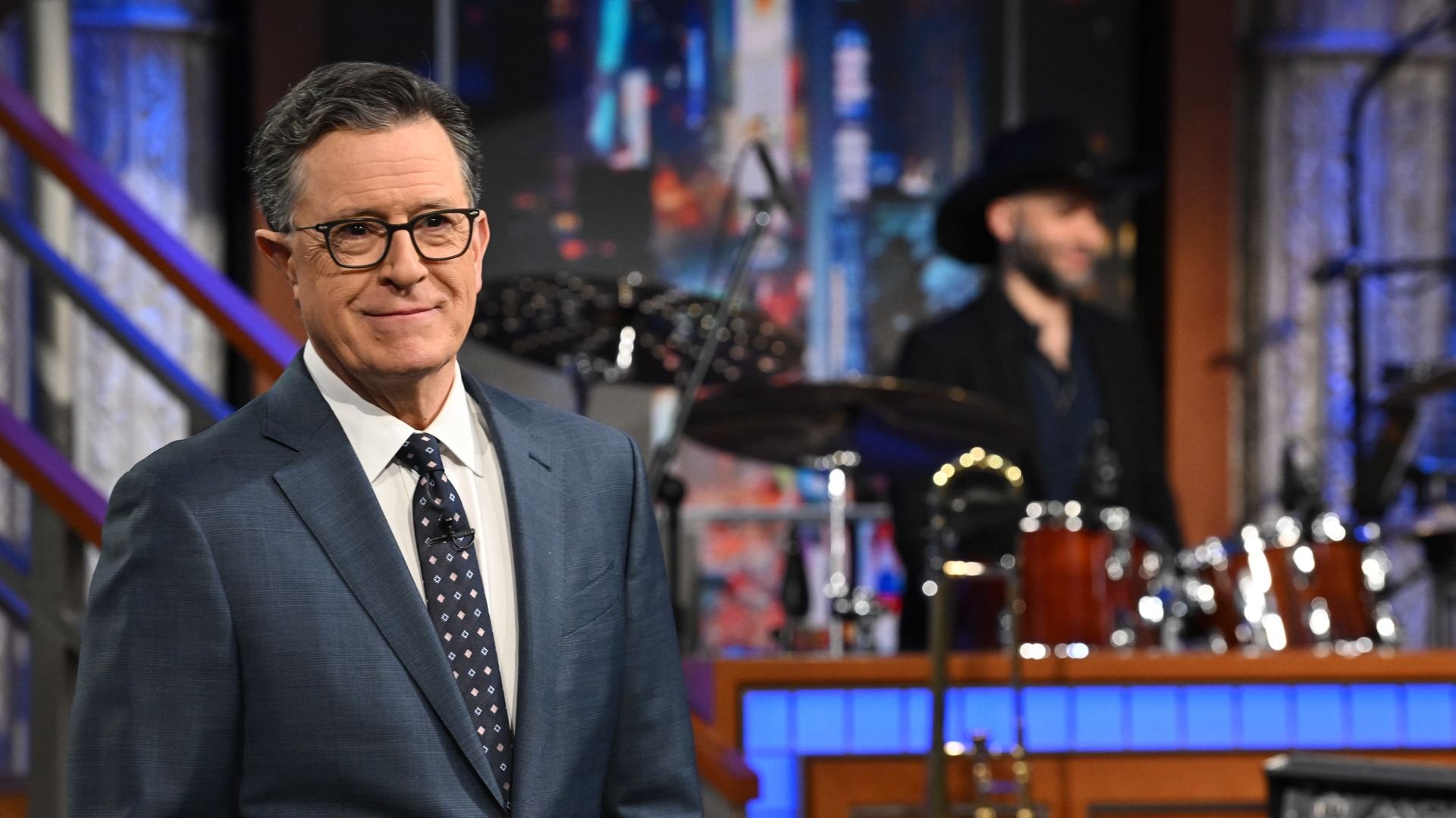 Stephen Colbert in a blue suit and patterned tie standing in front of a stage with musical instruments and a musician blurred in the background.