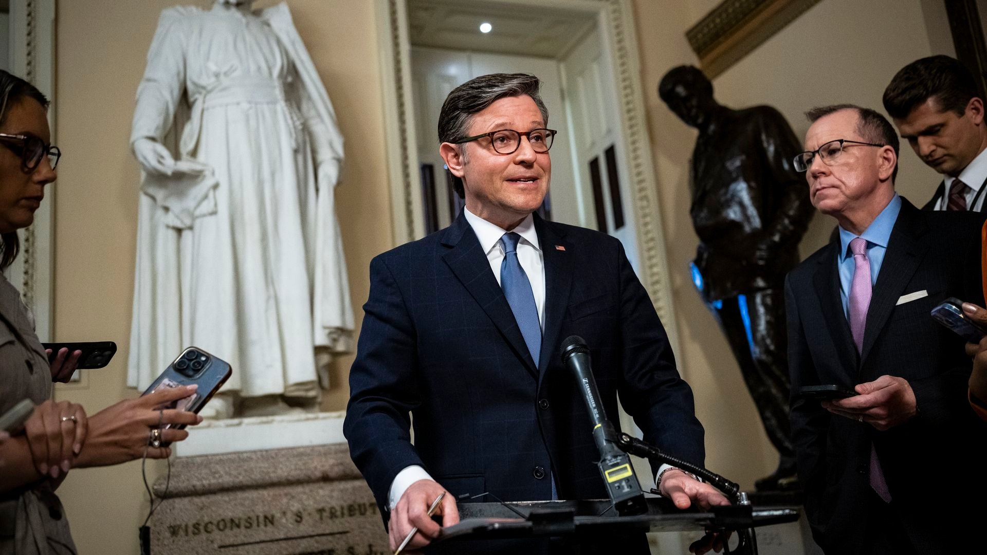 Politician in a navy suit and blue tie speaks at a podium as reporters hold phones; a white statue and doorway surround him in a marble-walled hallway.