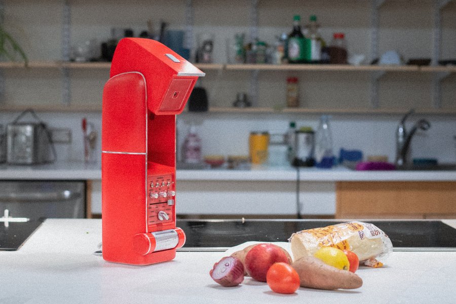 On a counter, a red Kitchen Cosmo device points its camera at assorted vegetables