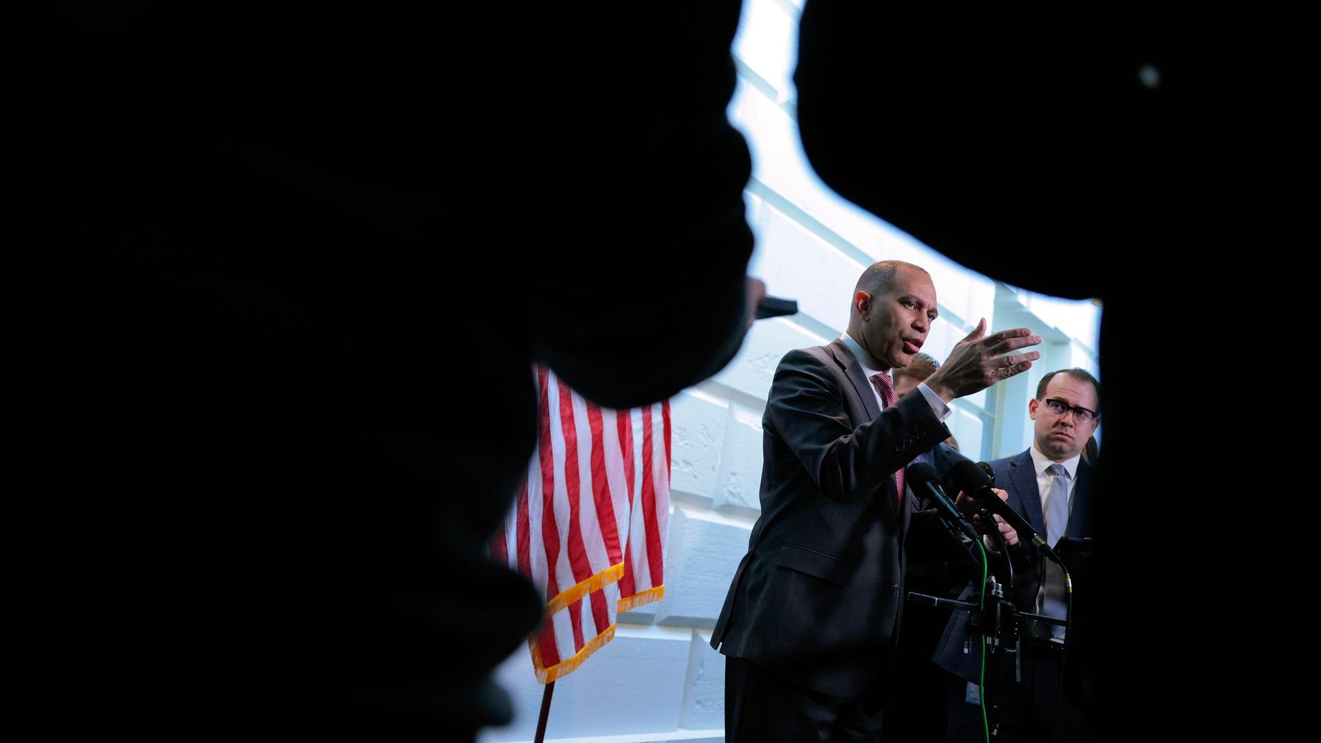 Man in suit speaking at a podium with microphones, accompanied by another man in glasses, framed by silhouettes, with a U.S. flag in the background against a white wall.