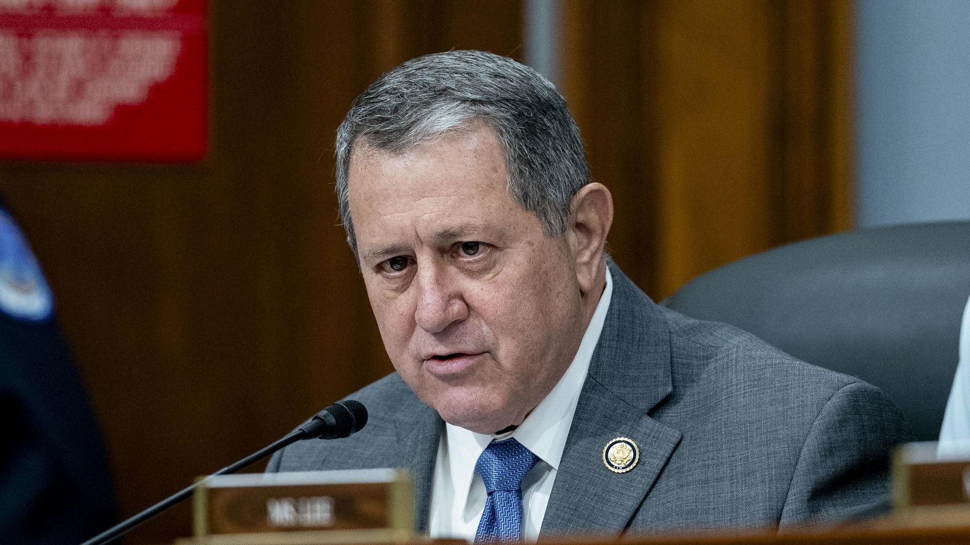 A man in a gray suit and blue tie sits at a wooden desk with a microphone, a circular lapel pin, and a serious expression during a formal hearing.