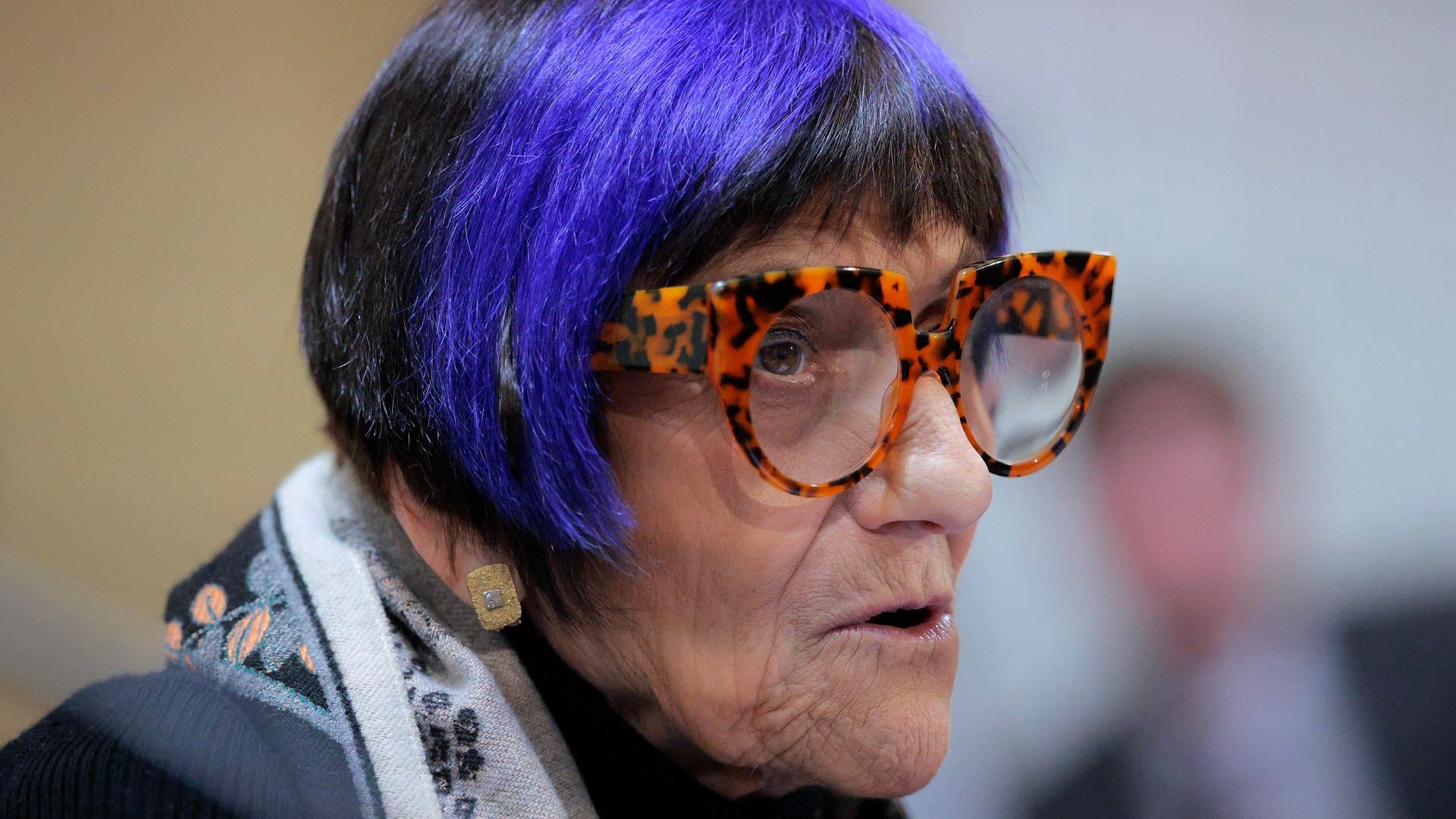 A woman with short black and purple hair, wearing large tortoiseshell glasses and a patterned scarf, speaking or in mid-conversation with a neutral background.
