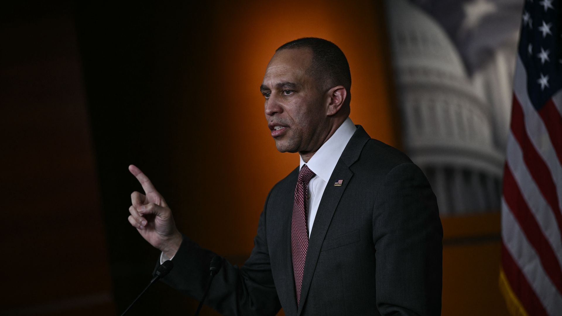 A man in a dark suit and red tie gestures with his right hand while speaking in front of microphones, with an American flag and a blurred government building backdrop behind him.