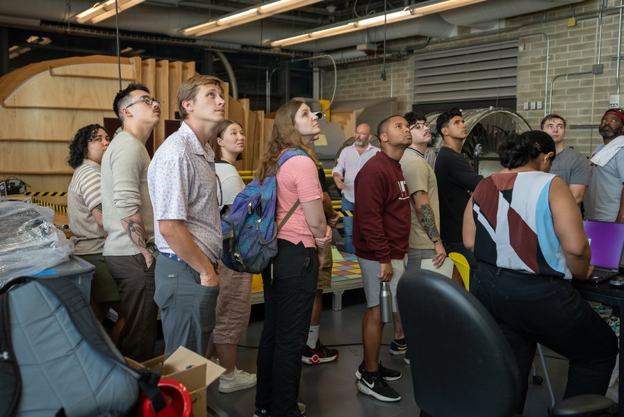 A group of about a dozen students standing in a lab, most of them looking at something out of frame