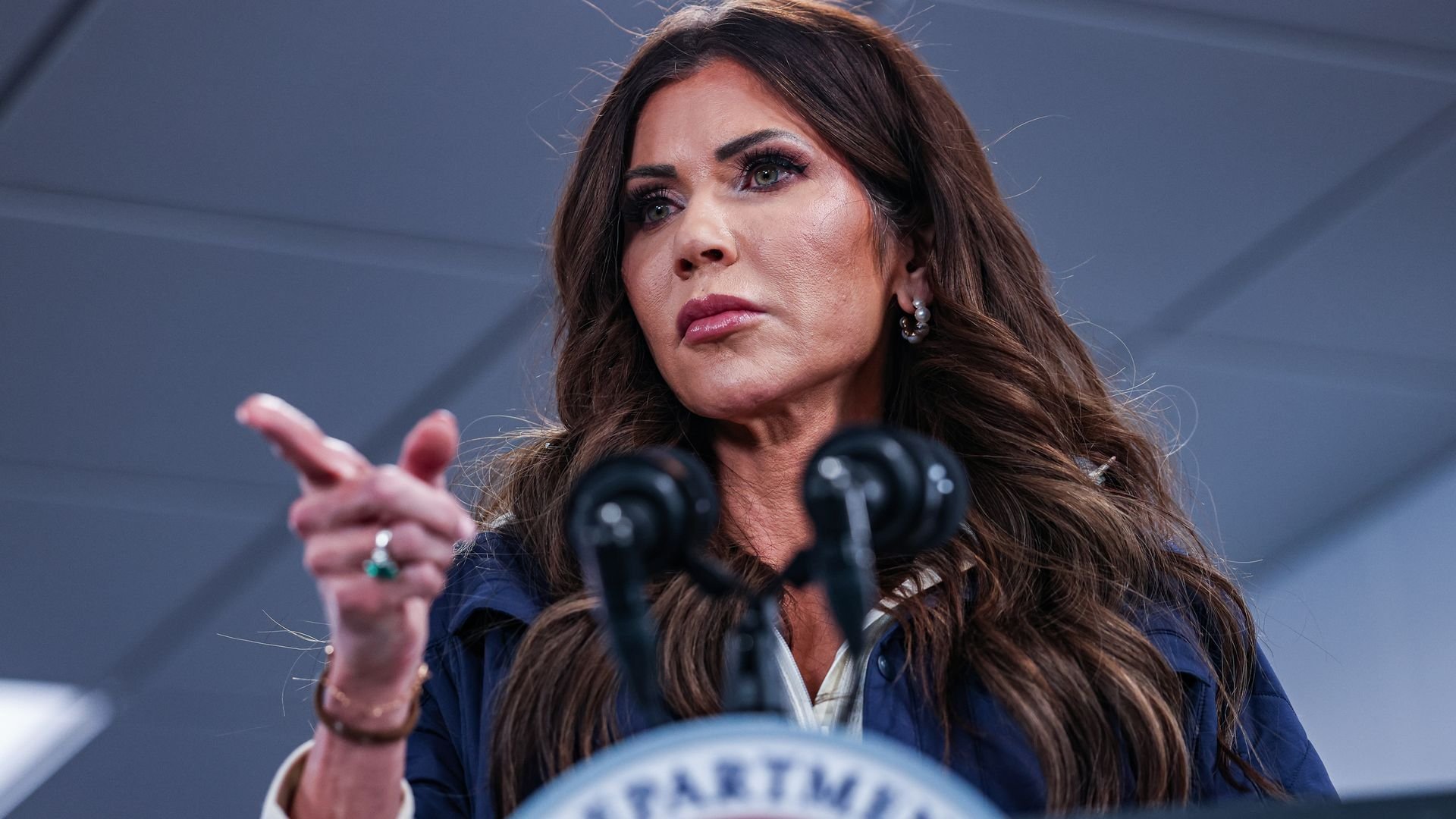 Noem wearing a blue zip-up over a white shirt standing at a podium and pointing her finger into the crowd with a straight expression
