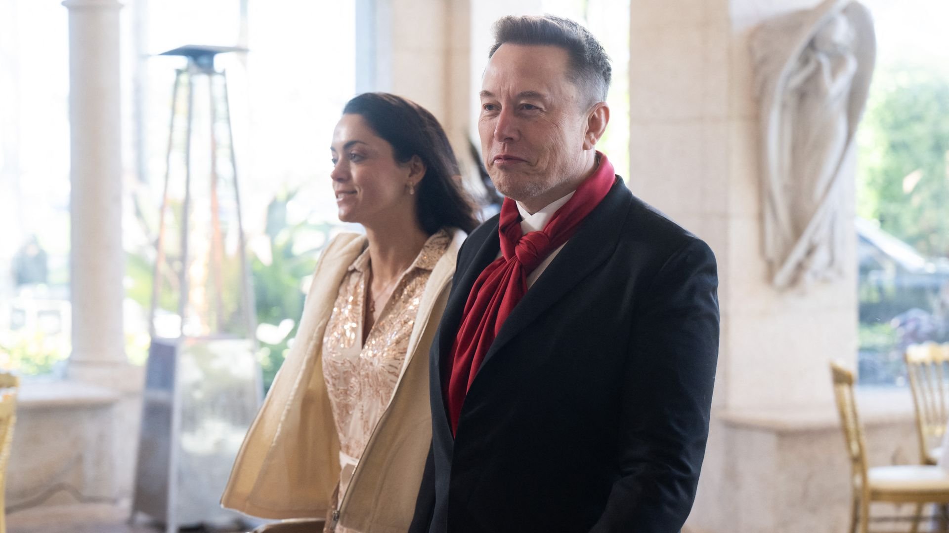 Elon Musk — wearing a red scarf, a white collared shirt and a black jacket — walks into an event at Mar-a-Lago alongside a brunette woman wearing gold.