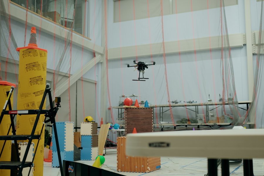 A drone flying an indoor obstacle course with barriers, ramps, and colorful cones in a large, well-lit room.