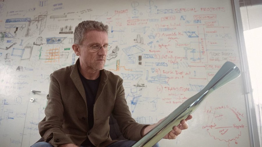 Carlo Rattis holds his Olympic torch, a metal cylinder with a flared end. Behind him is a whiteboard with equations and diagrams.