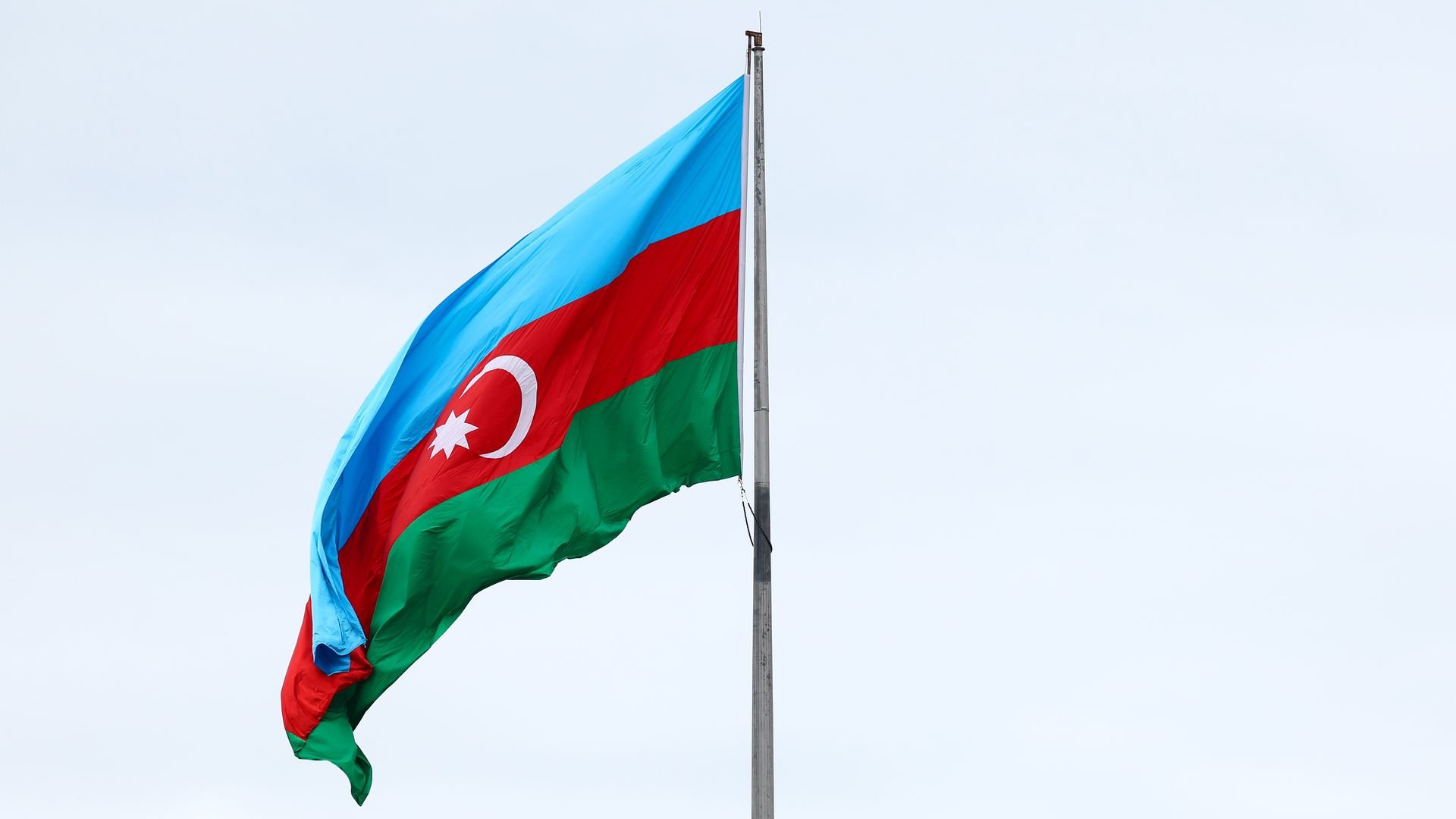 A flag with horizontal light blue, red, and green stripes, featuring a white crescent and an eight-pointed star in the center of the red stripe, flying on a metal pole against a cloudy sky.
