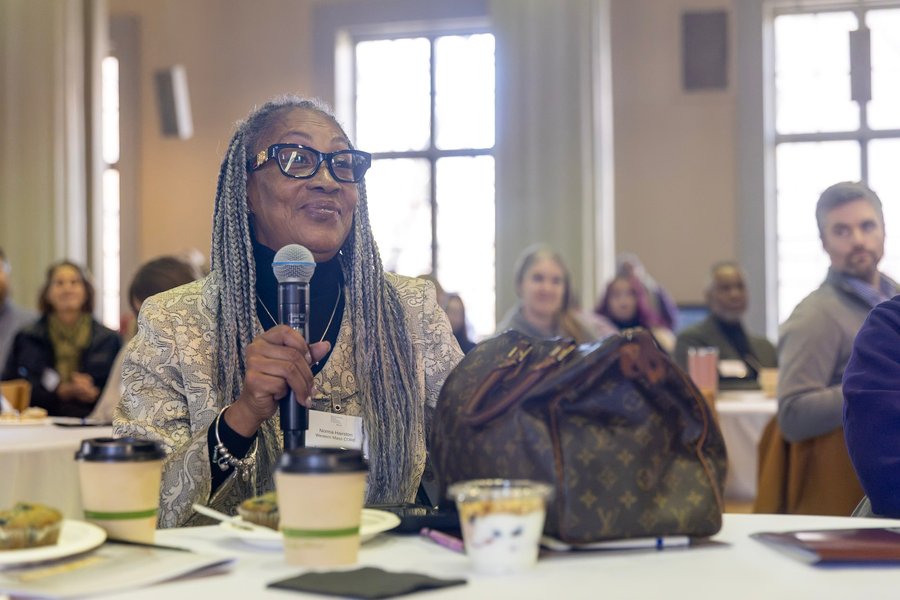 A woman with long dreadlocks seated at a table with others at tables behind her speaks into a microphone