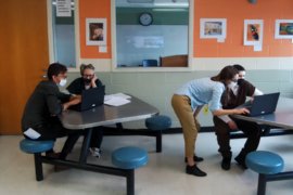 Four people share two laptops at separate tables in a tidy cinderblock room. 