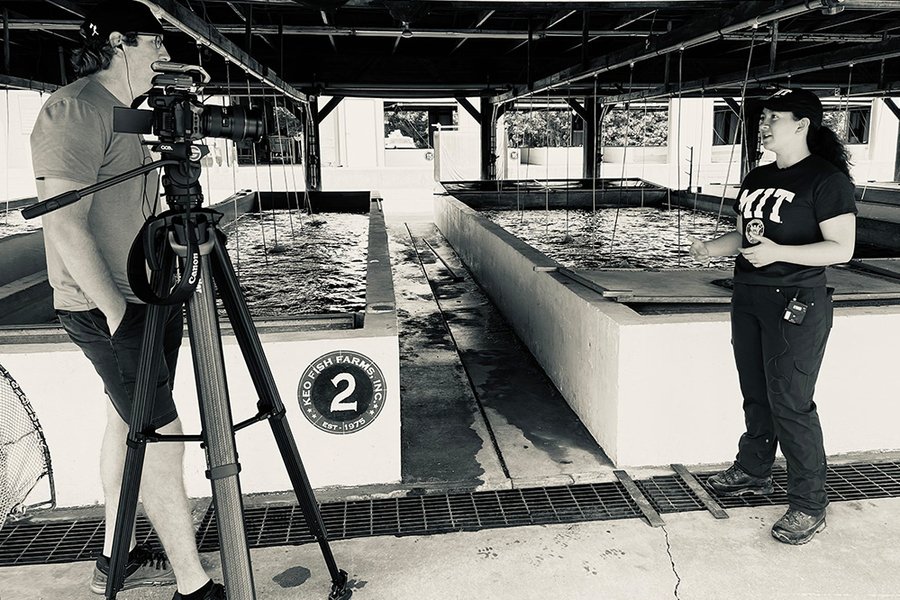 Grayscale photo of a man with a camera on a tripod talking to Kik Hayano, who stands before two raised pools.