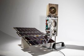 Photo of a small satellite against a white backdrop. The satellite has solar cells toward the bottom and a small boom with a gold cube at the top.