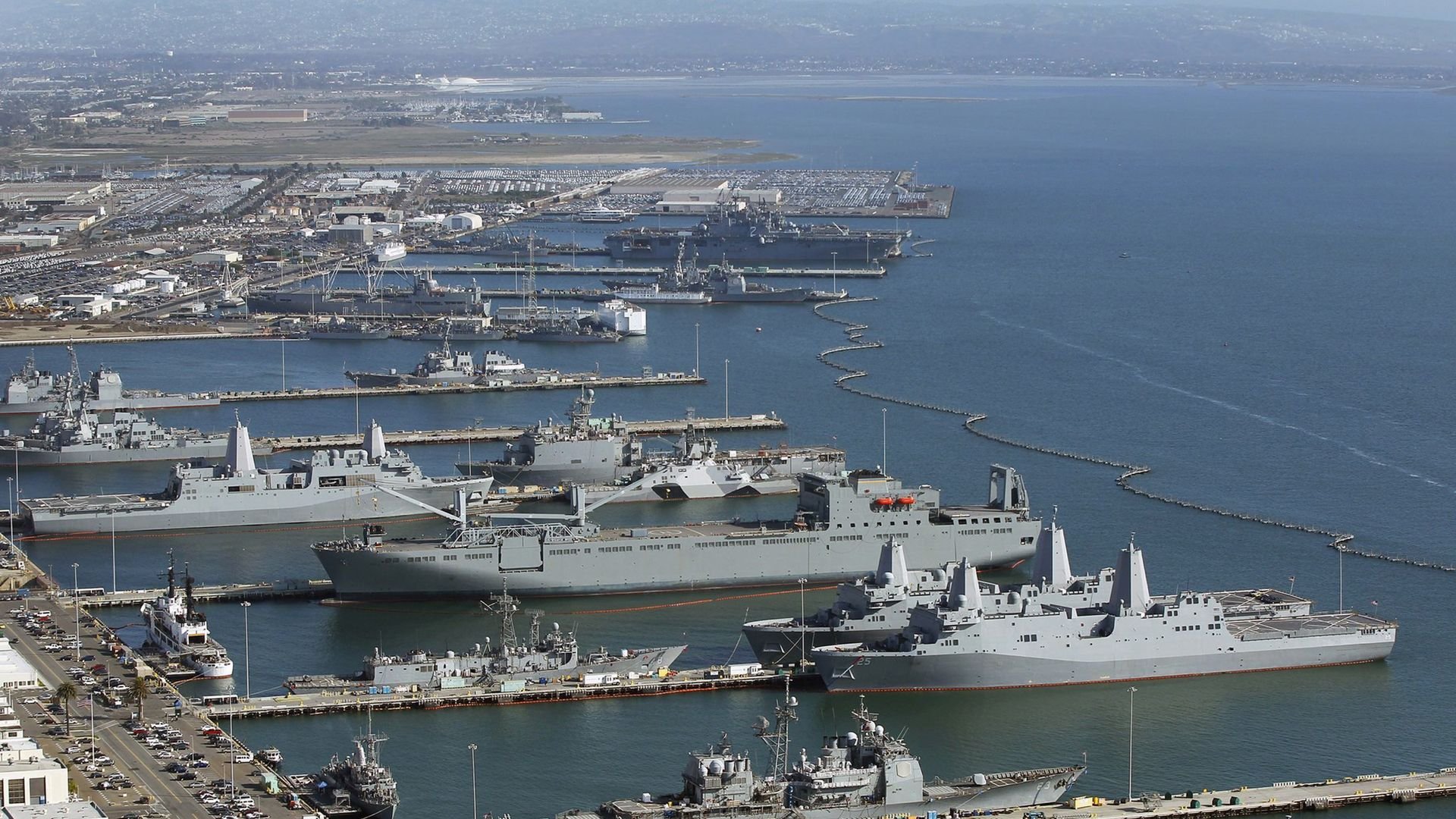 Aerial view of the large Naval Base San Diego, with multiple gray military ships docked along several piers extending into calm blue water under a clear sky.
