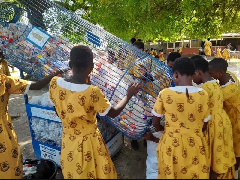 High school students in yellow uniform dresses handle a large wire basket filled with plastic waste