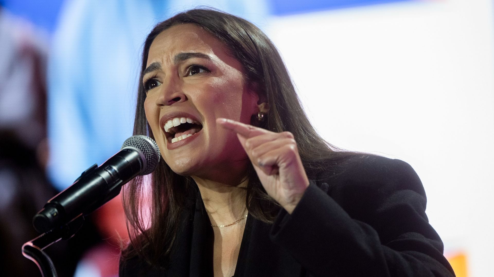 A woman with long dark hair wearing a black jacket speaks passionately into a microphone, with her mouth open and finger raised, against a blurred colorful background.