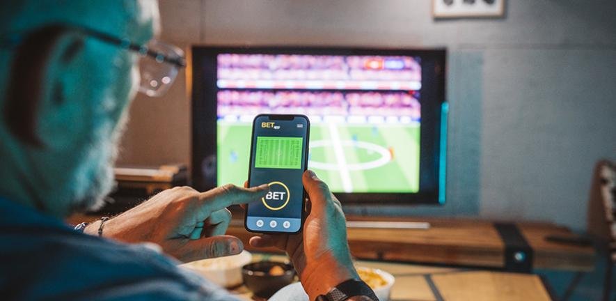 Man sitting on sofa at home and watching a football match. He is using smartphone for sports betting