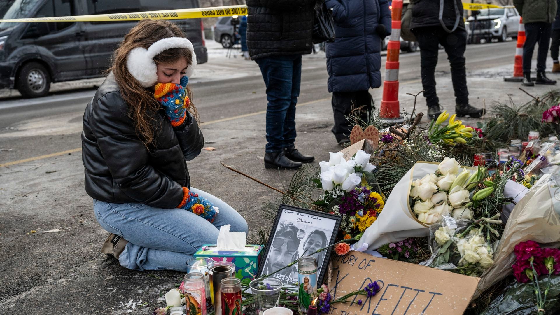 A woman wearing white earmuffs and colorful mittens kneels at a memorial for Alex Pretti made of candles and flowers.