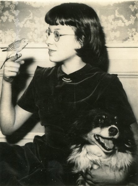 Sepia toned image of a young Harriet Latham with a parakeet perched on her right index finger, and her left arm around a happy-looking dog