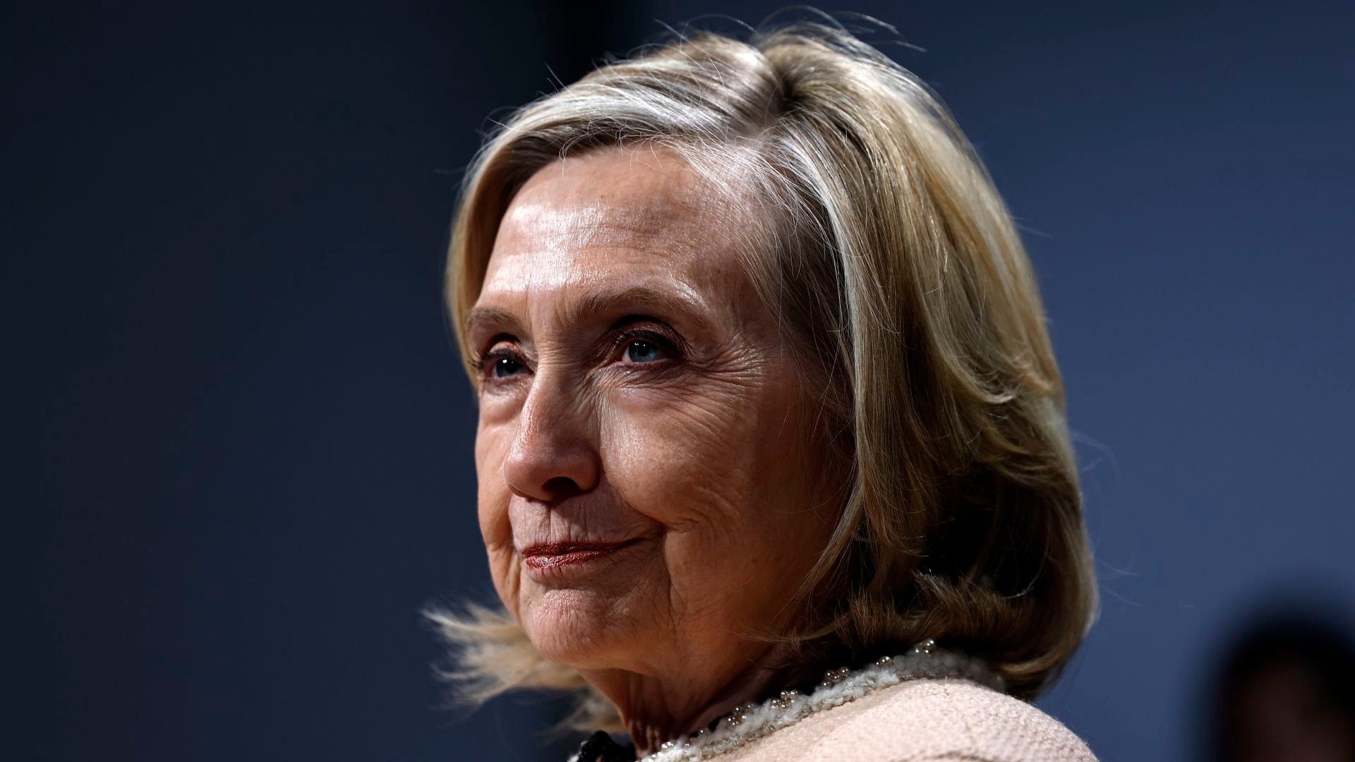Close-up of a woman with blonde hair and blue eyes, wearing a light-colored top with a pearl necklace, against a dark blurred background.