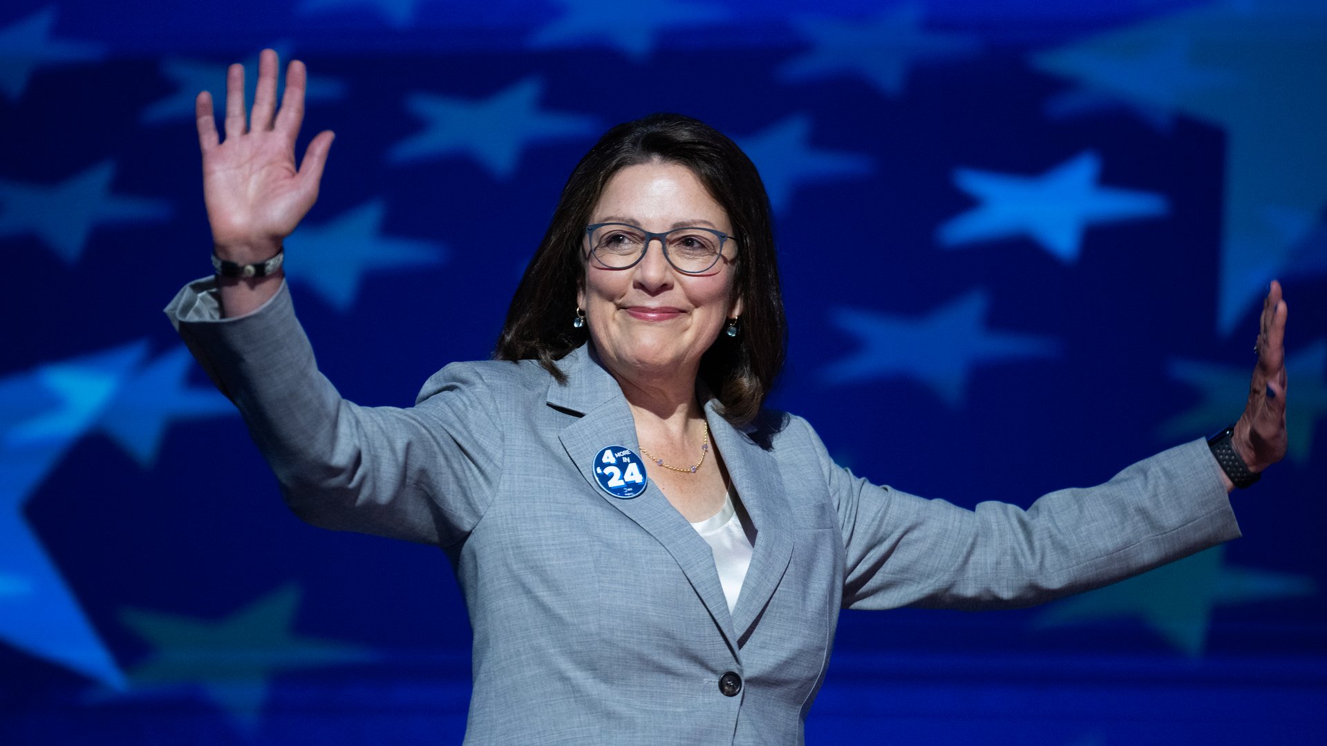 Woman in gray blazer and glasses waving with both hands, smiling, with a blue background featuring large white stars and a 