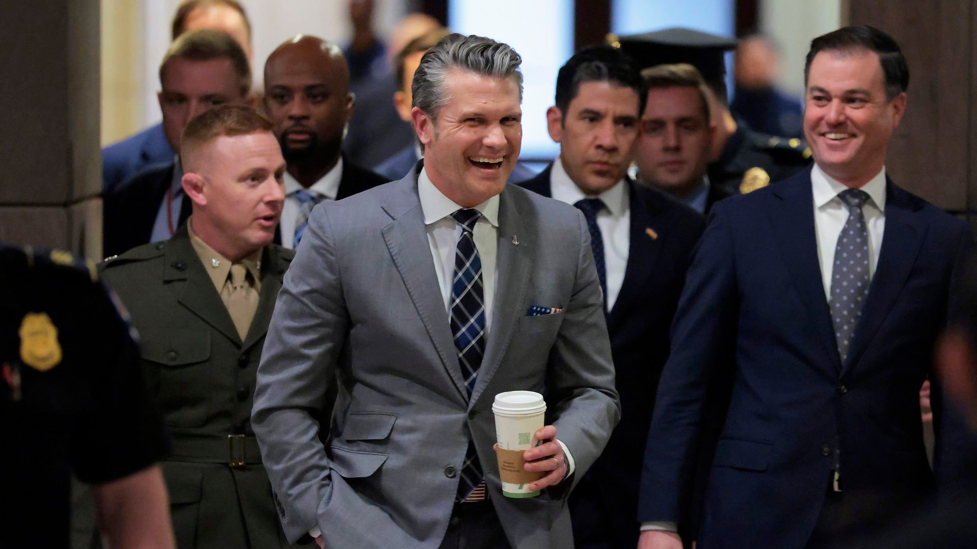 A group of men in suits and uniforms walking together indoors, two men in front smiling, one holding a coffee cup, the other carrying a briefcase.