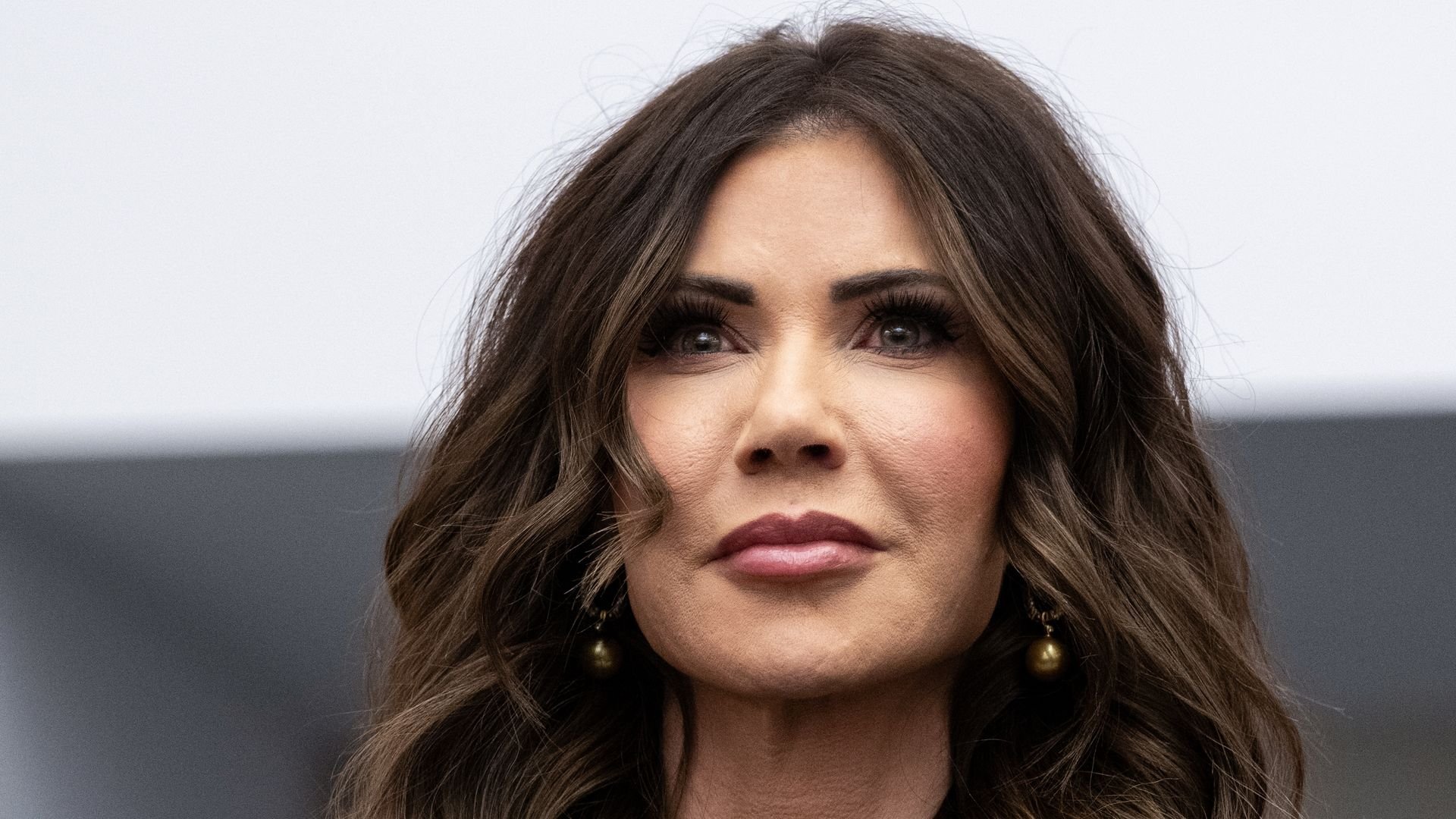 Close-up of a woman with long wavy brown hair, dark eyes and wearing dark round earrings against a blurry gray and white background.